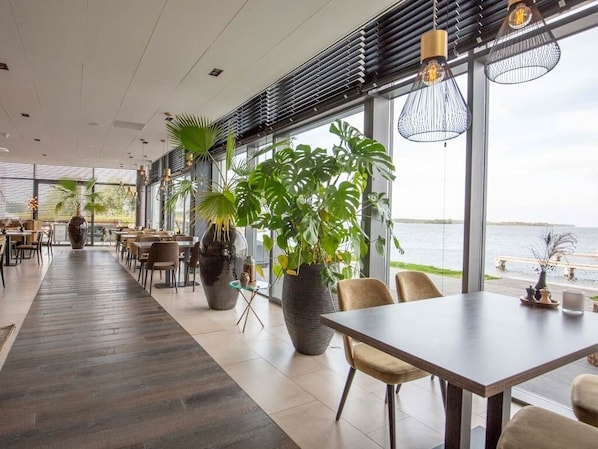 A modern restaurant with large windows, indoor plants, and a view of the water.