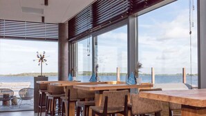 Un área de bar moderna con mesas y sillas de madera, ventanas amplias que ofrecen vista al agua y una escultura decorativa con forma de viento.