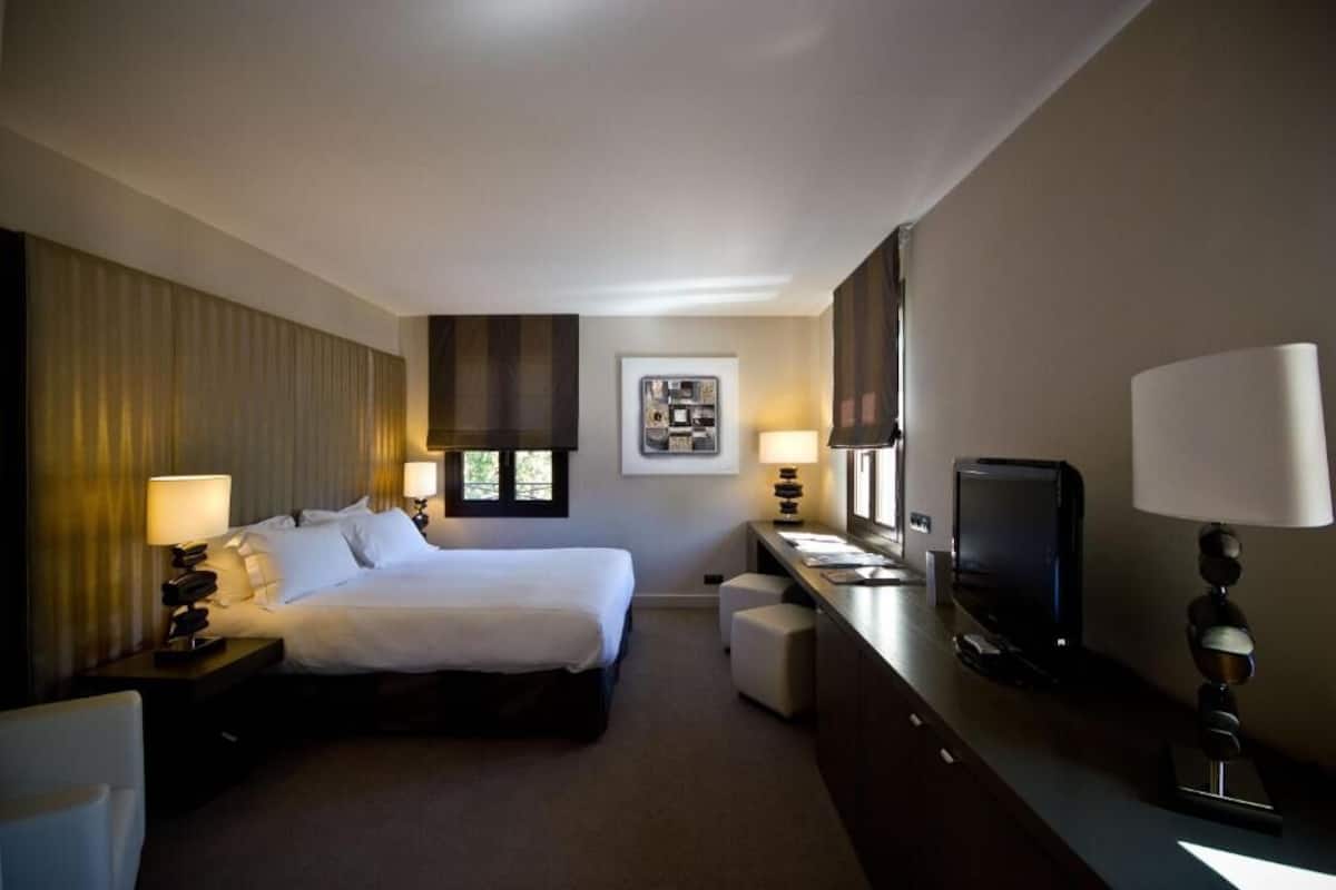 Quarto de hotel com cama, escrivaninha com televisão, uma cadeira e uma pequena mesa com abajur.