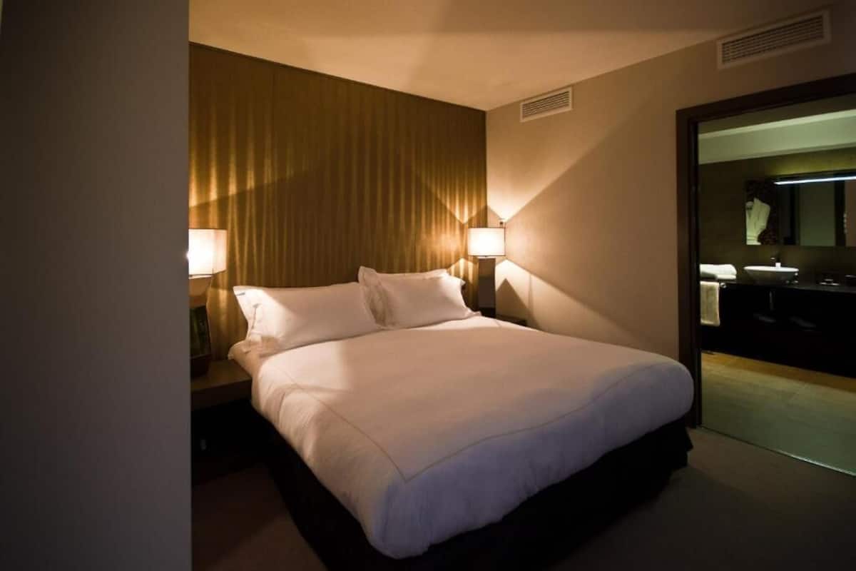 Quarto de hotel com cama, duas luminárias de cabeceira, espelho e janela com cortinas.