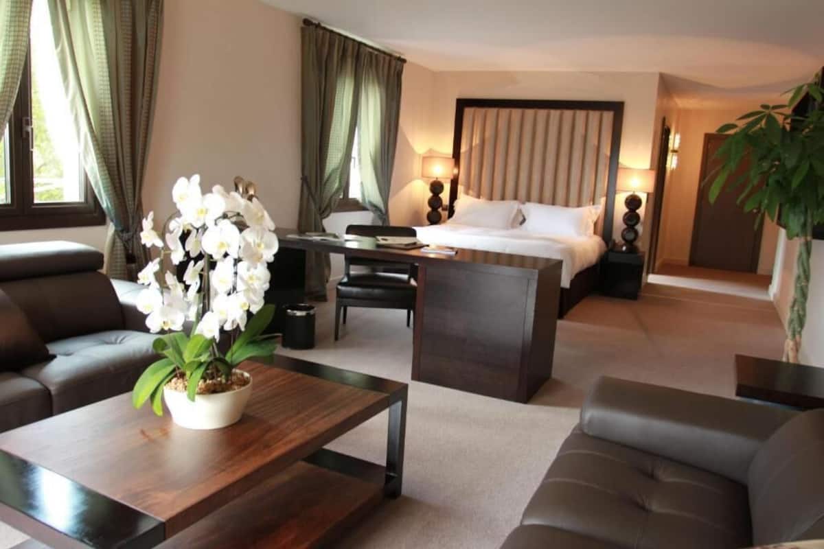 Quarto de hotel com cama, escrivaninha, duas poltronas, mesa de centro e planta em vaso.