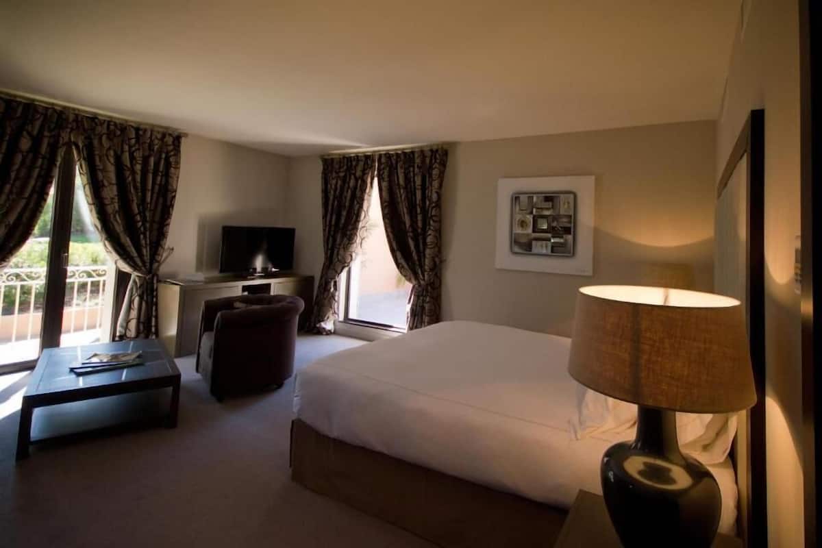 Quarto de hotel com cama, escrivaninha, televisão, luminária e janela com cortinas.