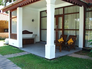A covered outdoor seating area with a bench and two chairs, a stone wall, and a large window with curtains.