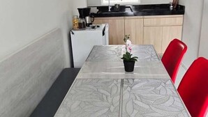 A dining area with a table, chairs, a small potted plant, and a kitchen in the background. - Residencial New Stilo (Aracati)
