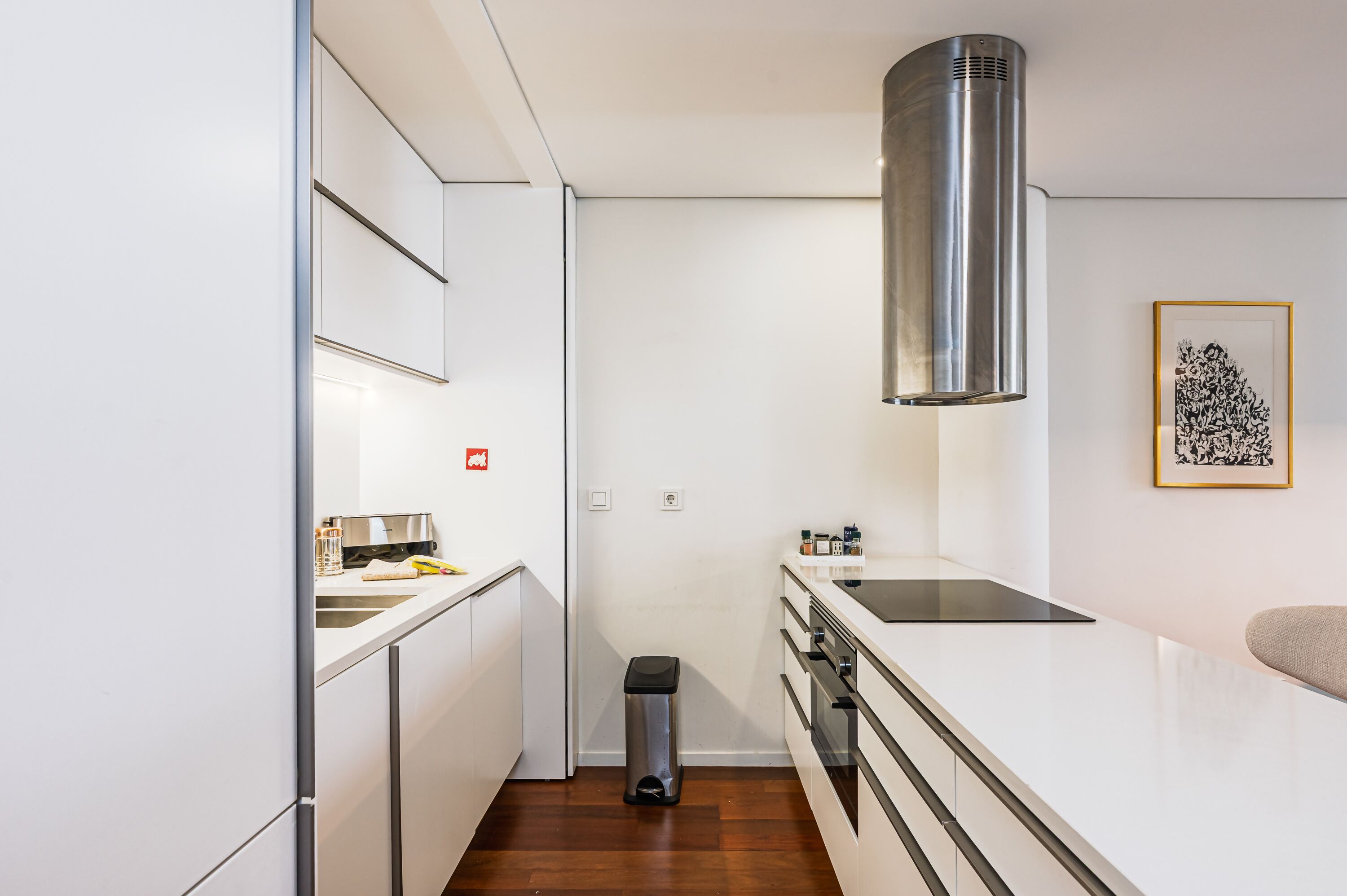 Una cocina moderna con armarios blancos, una campana extractora de acero inoxidable, una placa de inducción negra y un pequeño contenedor de basura.