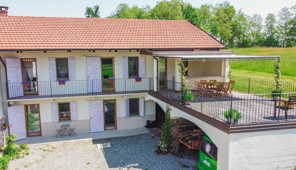 A two-story house with a red-tiled roof, a balcony with a table and chairs, and a gravel driveway.