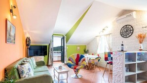 Ein Wohnzimmer mit einer Couch, einem Couchtisch, einem bunten Sessel, einem Esstisch mit Stühlen, einem Fernseher und einer Wanduhr.