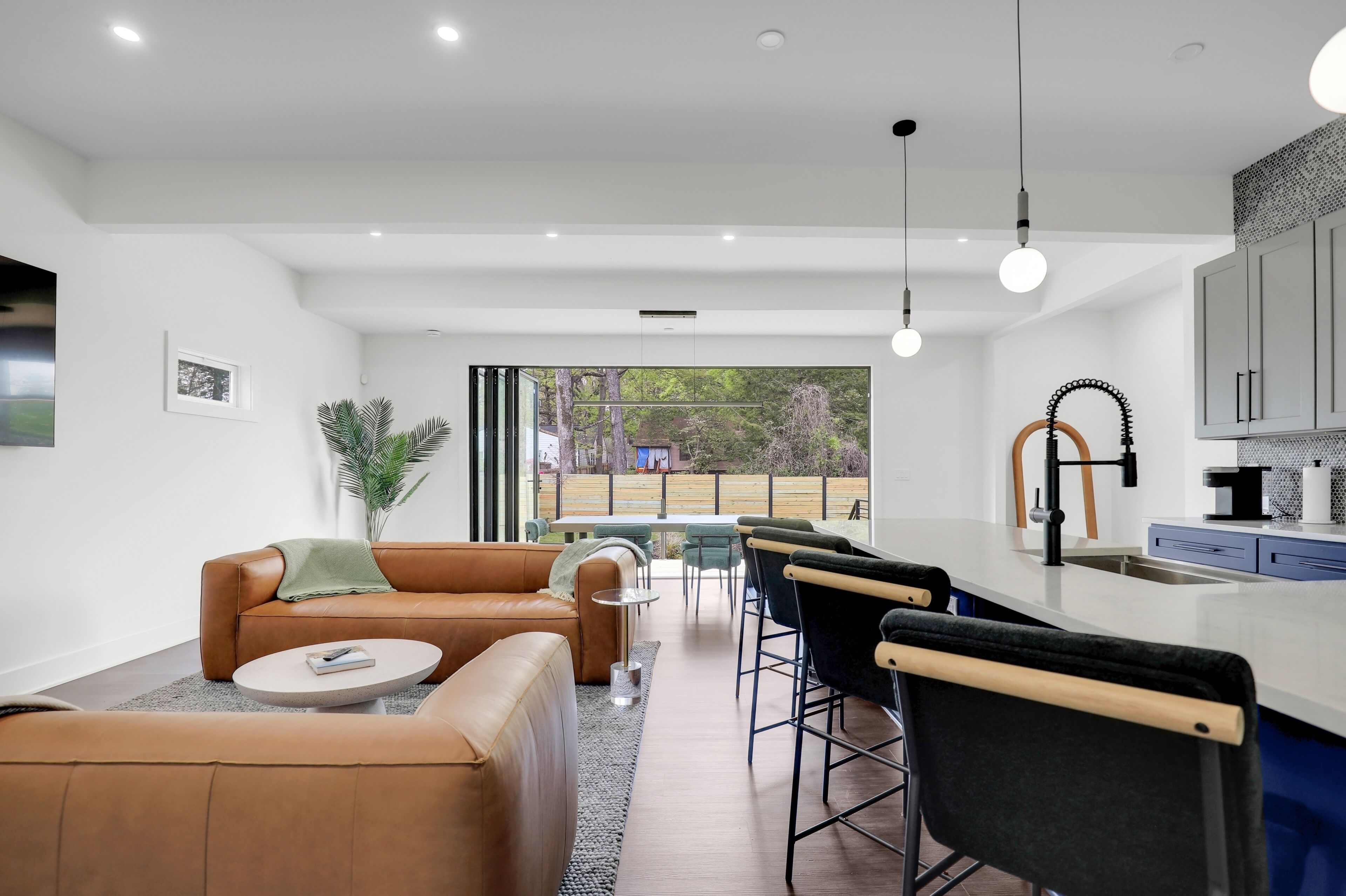 Een moderne woonkamer met een bank, salontafel en een eethoek met stoelen. Er is een keuken met een bar en een groot raam.