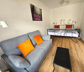 Un canapé gris avec des coussins orange, une table basse noire et un cadre de lit en métal avec un matelas blanc.