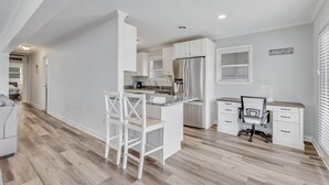 Una cocina moderna con armarios blancos, una isla integrada con taburetes de bar, un refrigerador y un comedor con un escritorio y una silla blancos.