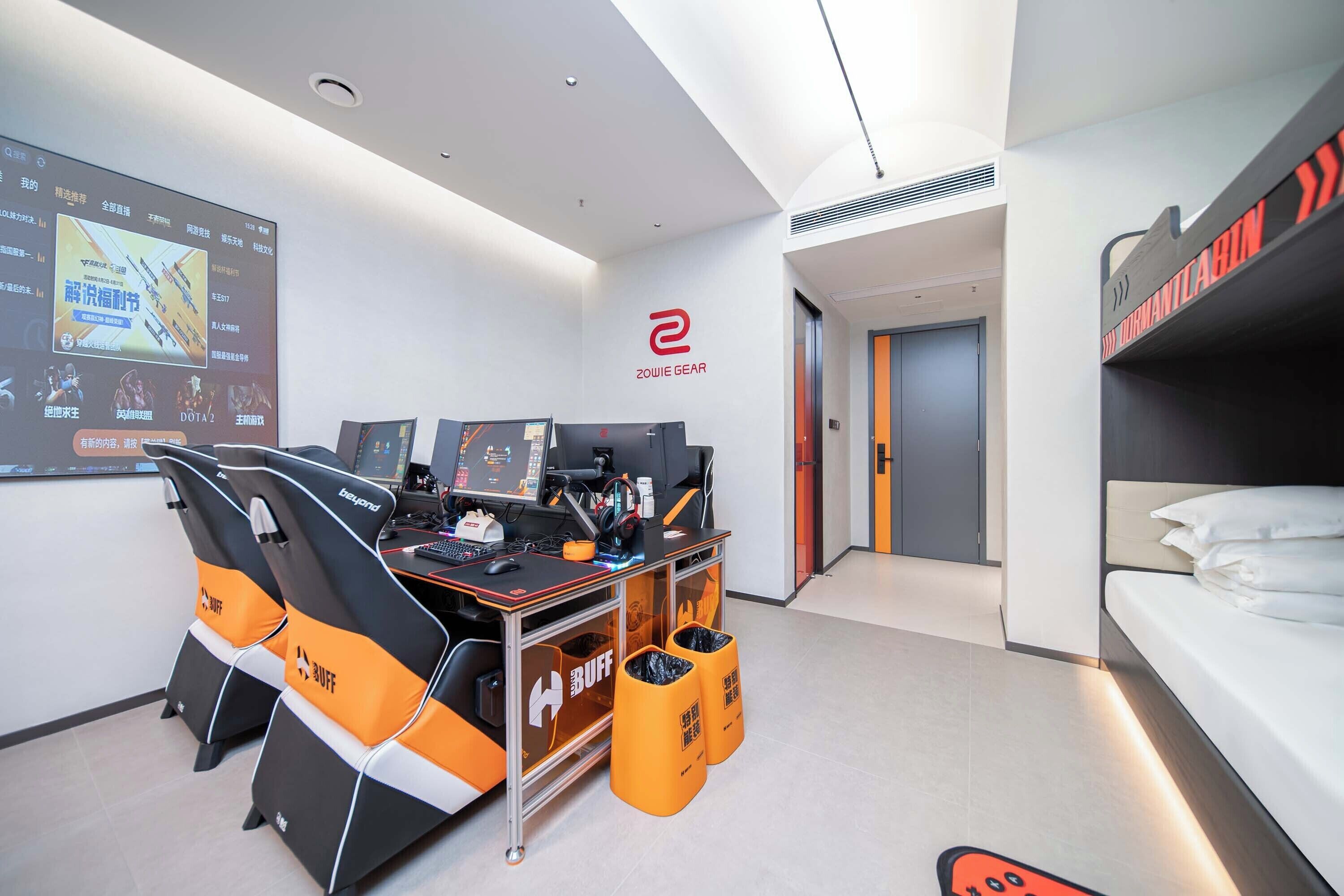A modern hotel room with a gaming setup, including two gaming chairs, a desk with a computer, and a large screen. There is a bunk bed with a sign on the wall and a trash bin.