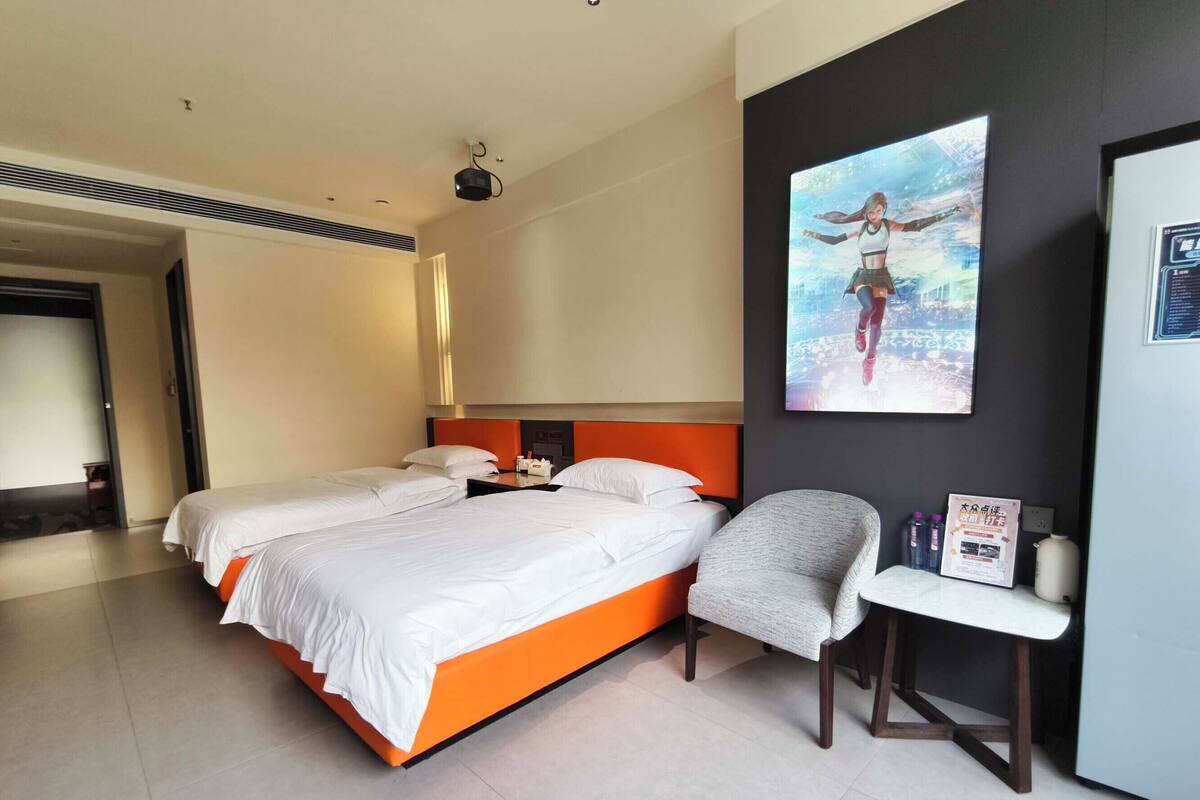 A hotel room with a double bed, a chair, a small table, a projector, and a poster on the wall.