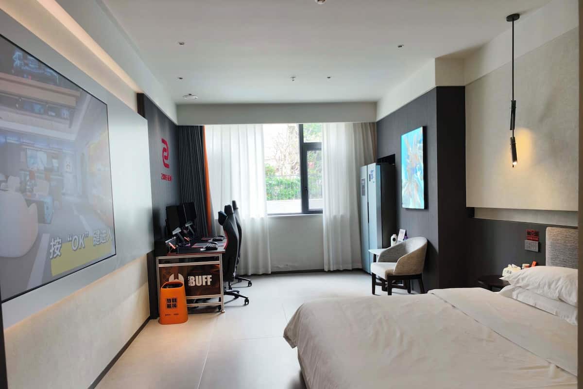 A modern hotel room with a large bed, a desk with a computer, a chair, a TV, and a window with curtains.
