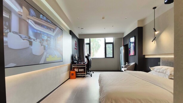 Une chambre d’hôtel moderne dotée d’un grand lit, d’un bureau avec un ordinateur, d’un écran de projection et d’une fenêtre donnant sur un paysage verdoyant.