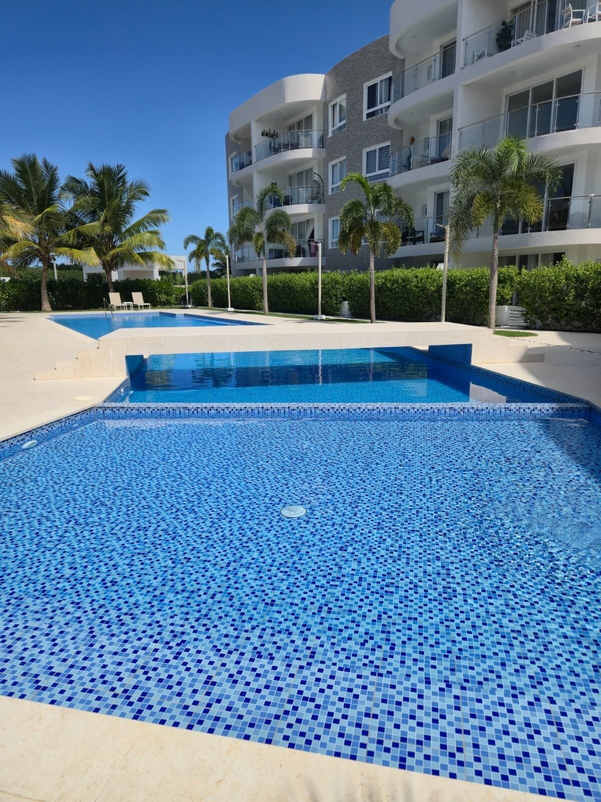 Una piscina con mosaicos azules, rodeada de palmeras y un edificio de varios pisos con balcones.