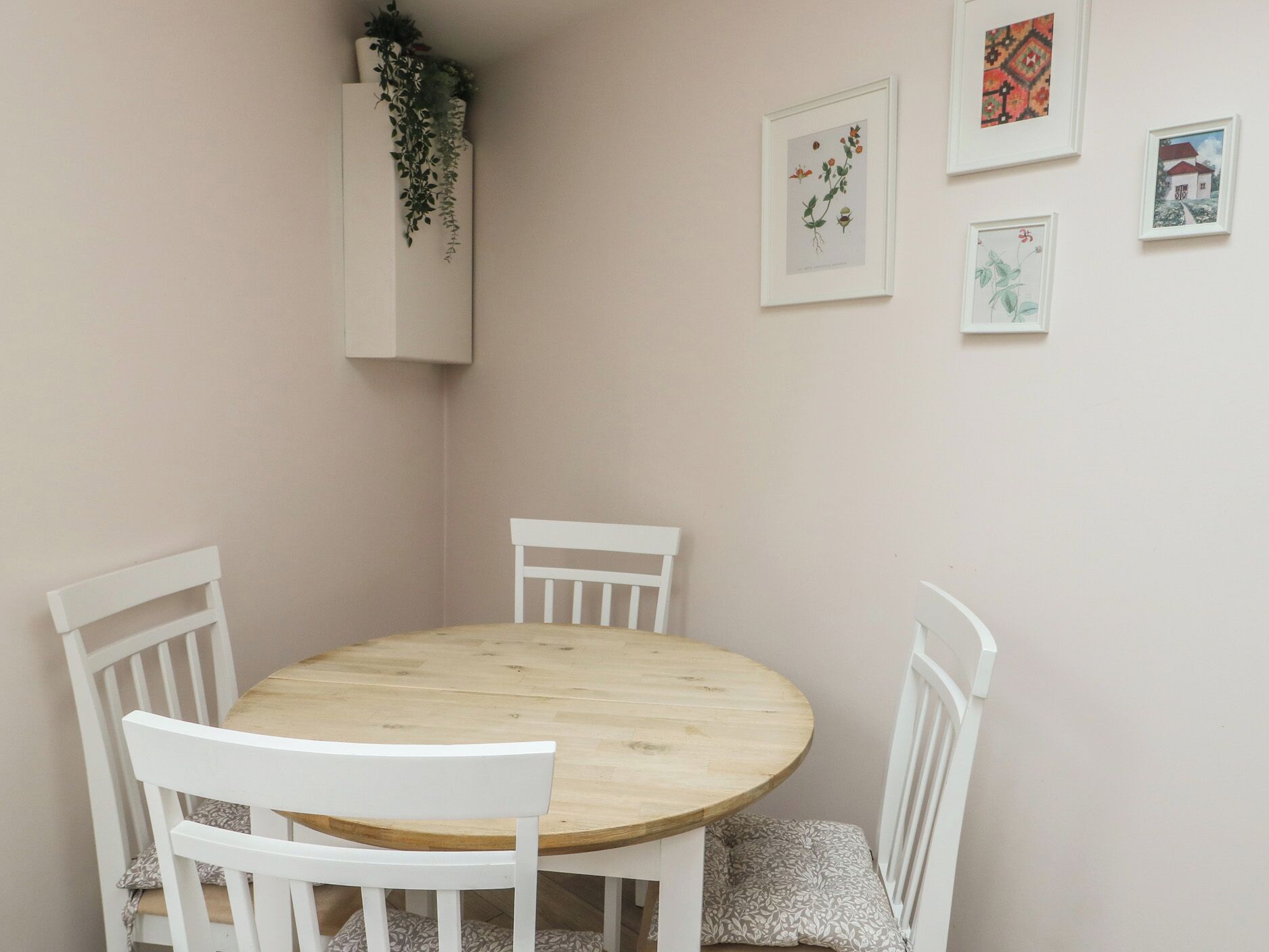Una mesa redonda de madera con cuatro sillas blancas, una pared blanca decorada con impresiones botánicas enmarcadas, y una maceta blanca con plantas verdes.