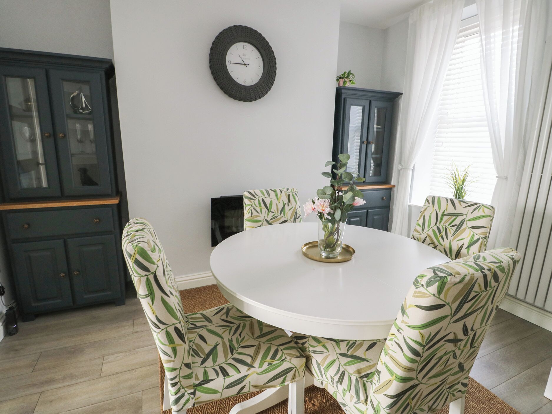 Een eethoek met een ronde tafel, stoelen met een bladmotief, een wandklok en donkere kasten.