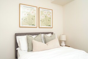 A bed with white bedding, multiple pillows, a nightstand with a lamp, and two framed botanical prints on the wall.