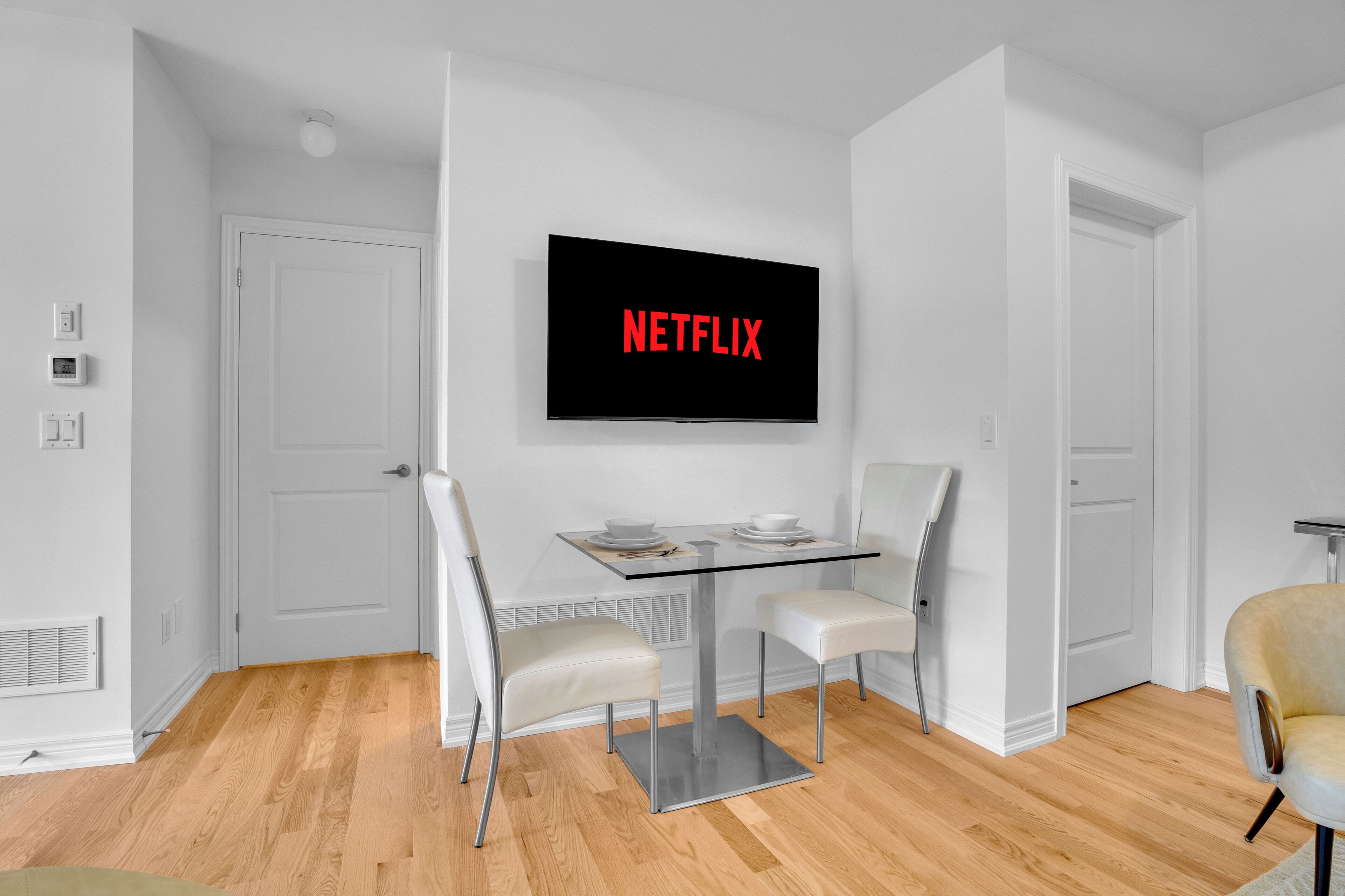 Un salon moderne avec une table basse en verre, deux chaises blanches, un téléviseur à écran plat fixé au mur qui affiche « NETFLIX », et un plancher en bois.