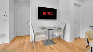 Un salón moderno con una mesa de centro de vidrio, dos sillas blancas, un televisor de pantalla plana montado en la pared mostrando 'NETFLIX' y un suelo de madera.