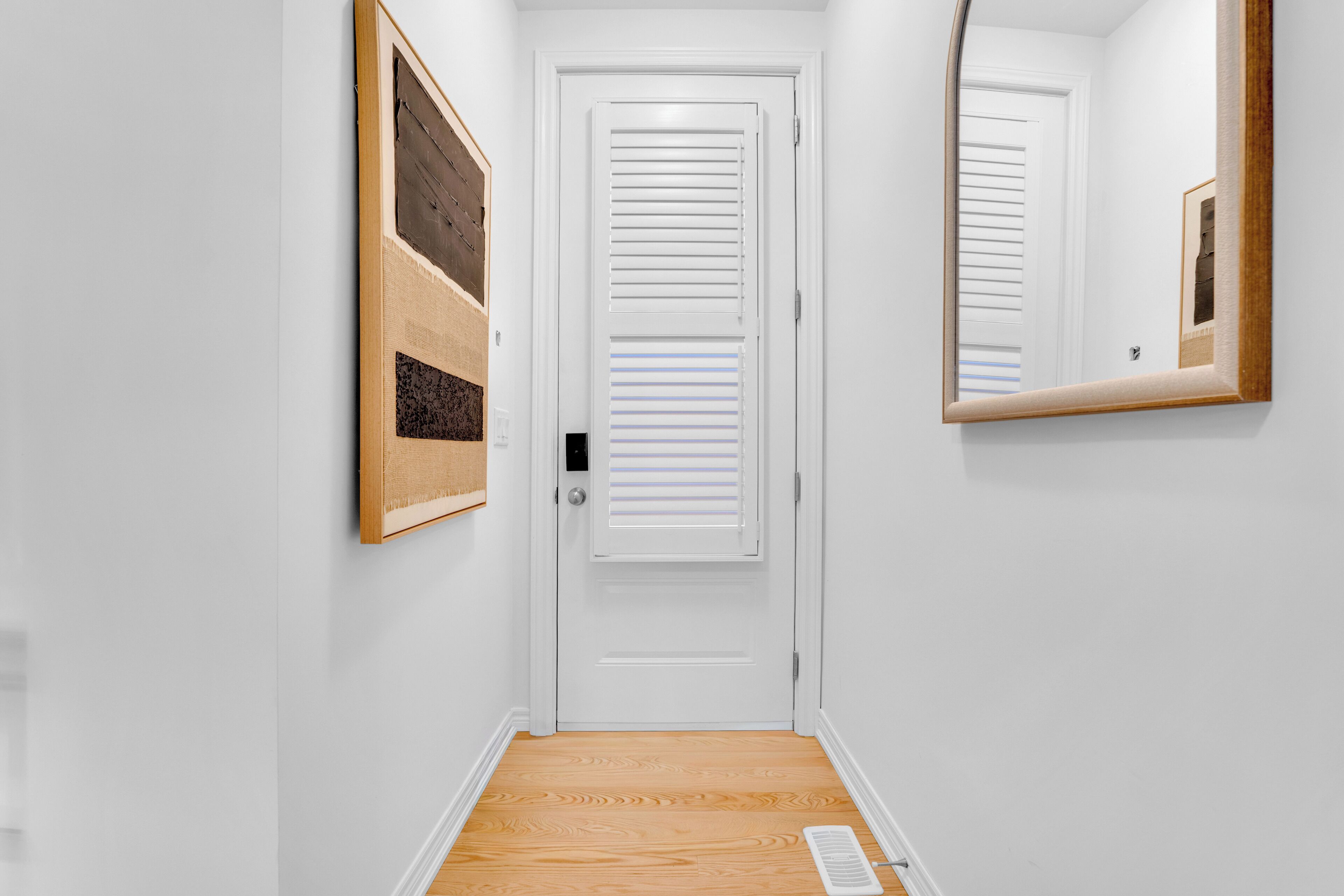 Un couloir avec une porte blanche, un miroir et une grille d’aération.