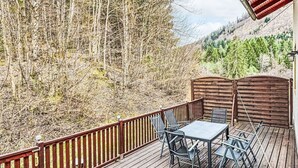 Une terrasse en bois, agrémentée d’une table et de chaises, entourée d’une rambarde en métal, et offrant une vue sur une zone boisée.