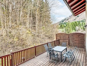 Eine Holzterrasse mit Tisch und Stühlen, umgeben von einem Metallgeländer, mit Blick auf eine bewaldete Gegend.