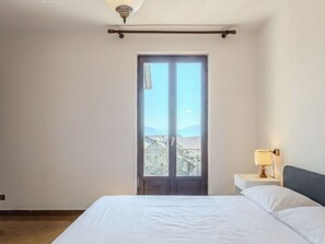 Et soveværelse med en seng, et natbord, en lampe og udsigt til en balkon med bjerge i baggrunden.
