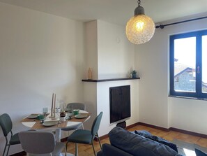 Et spiseområde med et bord dækket til fire personer, et fjernsyn ophængt på væggen og et stort vindue med udsigt til bygninger.