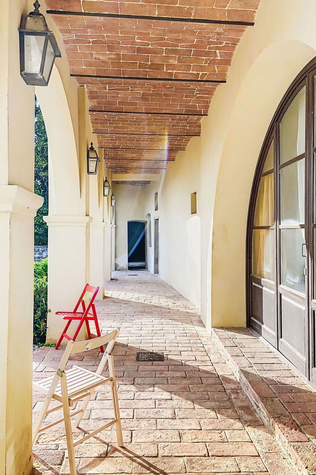 Un couloir couvert avec un sol en briques, une chaise en bois, une chaise rouge et une porte en verre.