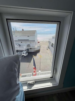 On voit, à travers une fenêtre, un bâtiment à la façade blanche, doté d’un balcon et devant lequel est garée une voiture.