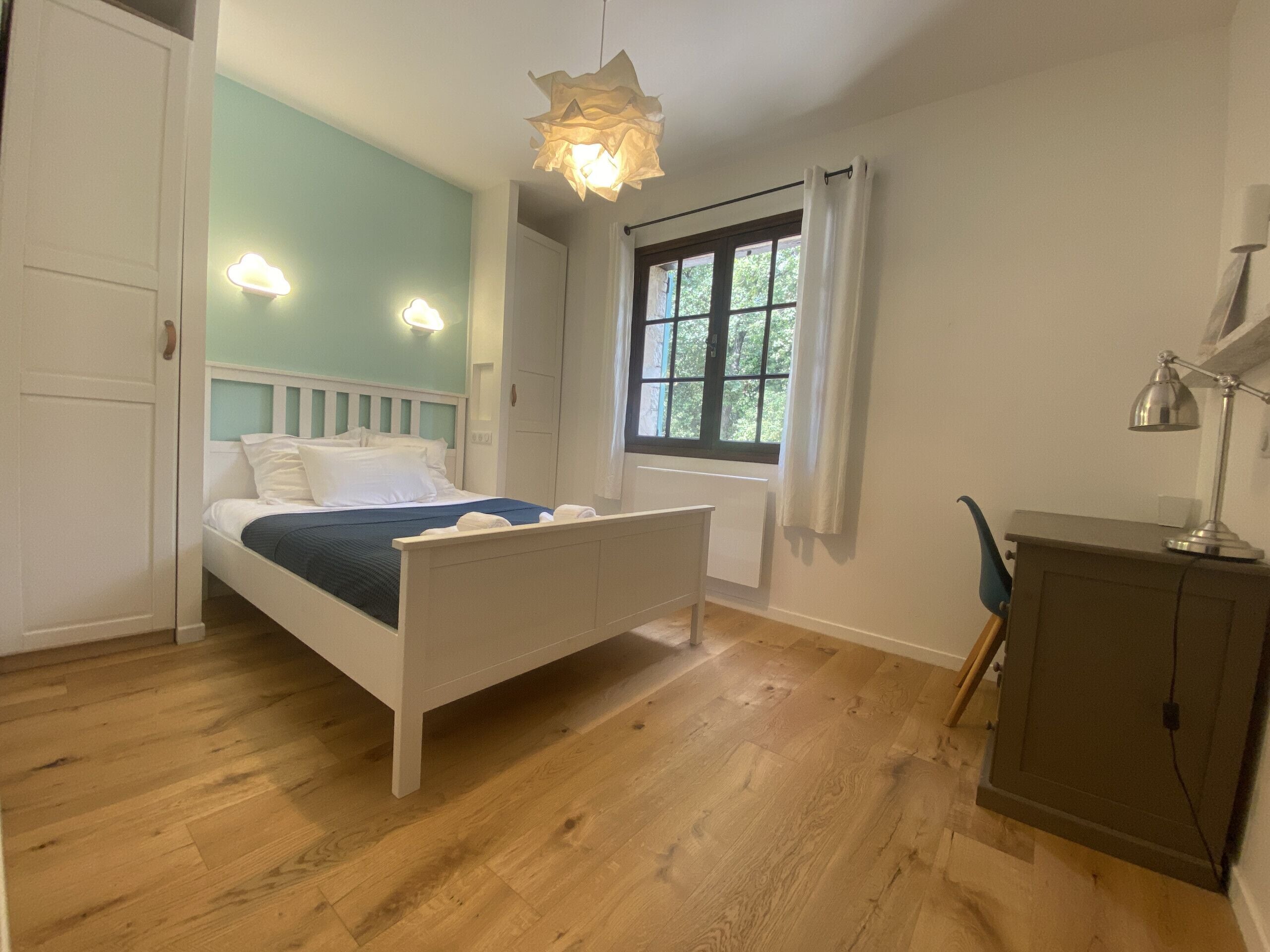 A bedroom with a bed, a desk, a chair, a window, and a hanging light fixture.