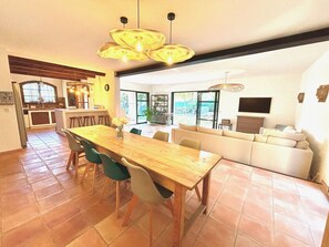 Un vaste espace de vie comprenant une table à manger en bois, des chaises vertes, un canapé beige et une grande fenêtre donnant sur l’extérieur.