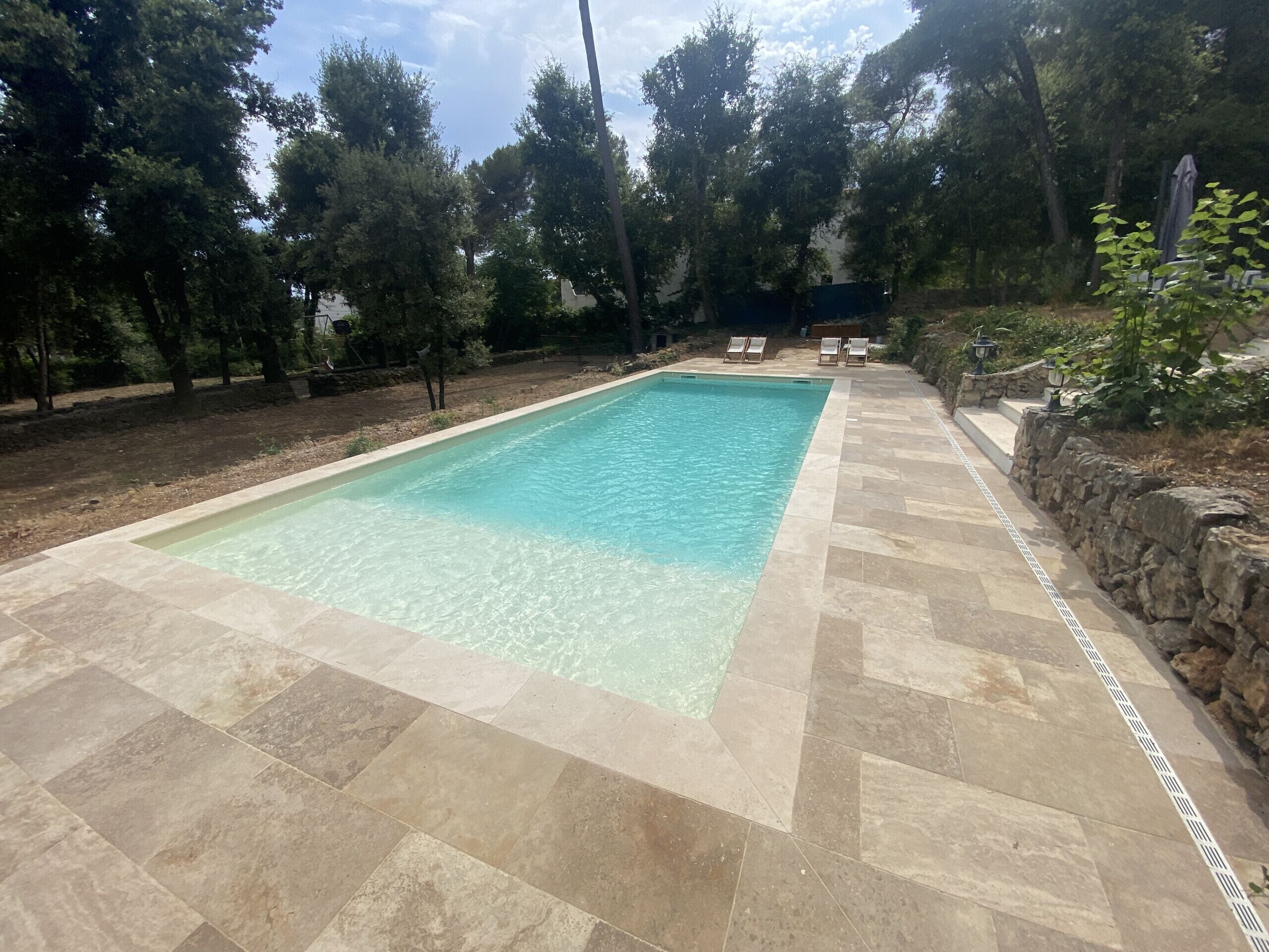 A rectangular swimming pool with clear water, surrounded by stone tiles and a stone wall.