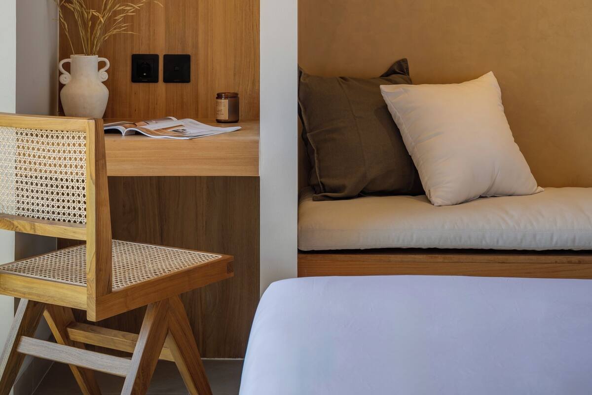 A room with a wooden desk, a wicker chair, a sofa with cushions, and a vase with plants.