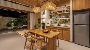 A modern kitchen with wooden cabinets, a stainless steel refrigerator, and a dining area with wicker chairs and a wooden table.