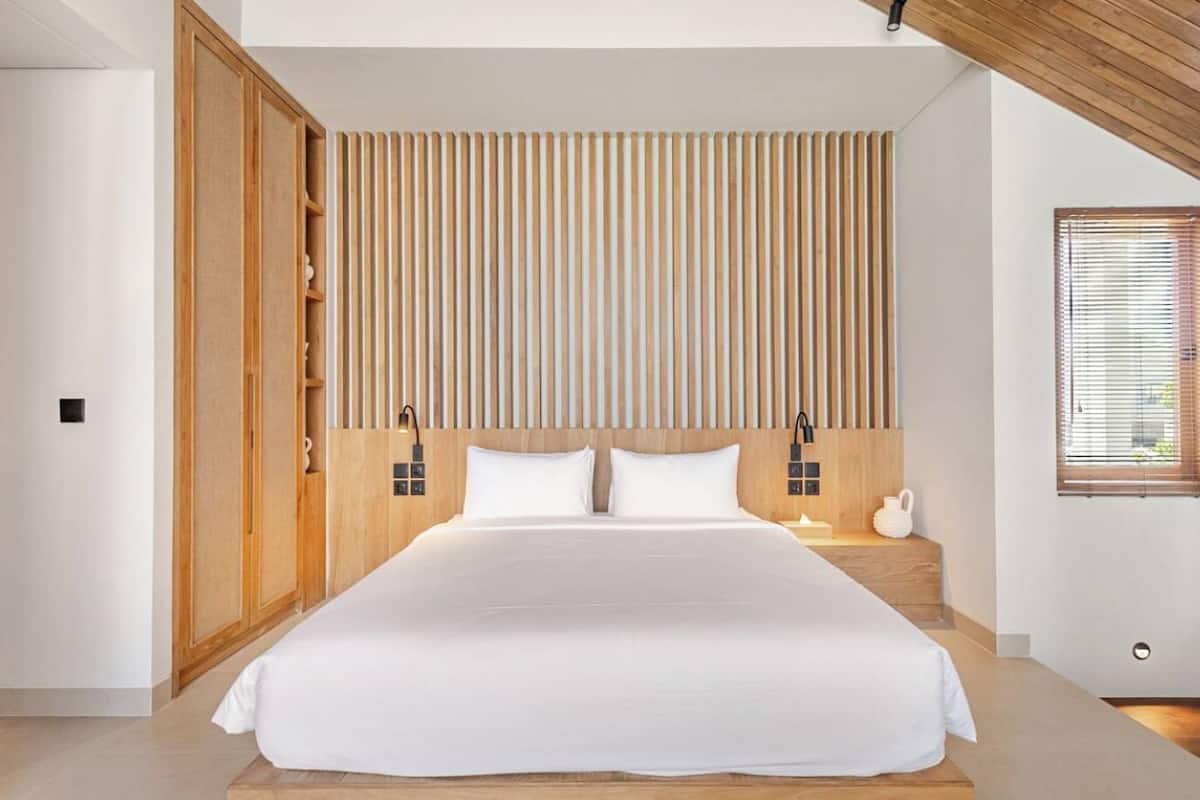 A modern bedroom with a large bed, wooden panel headboard, bedside lamps, and a window with blinds.