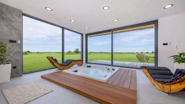A modern spa room with a wooden deck, a hot tub, and large windows offering a view of a green field. - Villa Aurora mit Sauna und Whirlpool (Dranske)