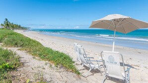 A beach with a white umbrella and a chair labeled 'NOMAR' on a sandy shore. - FlatCarneiros-Varanda com Vista-SH152 (Tamandaré)