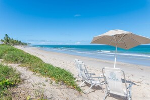 Ein Strand mit einem weißen Sonnenschirm und einem Stuhl mit der Aufschrift 'NOMAR' am sandigen Ufer.