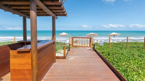 A wooden deck leading to a beach with umbrellas and a wooden structure. - FlatCarneiros-Varanda com Vista-SH152 (Tamandaré)