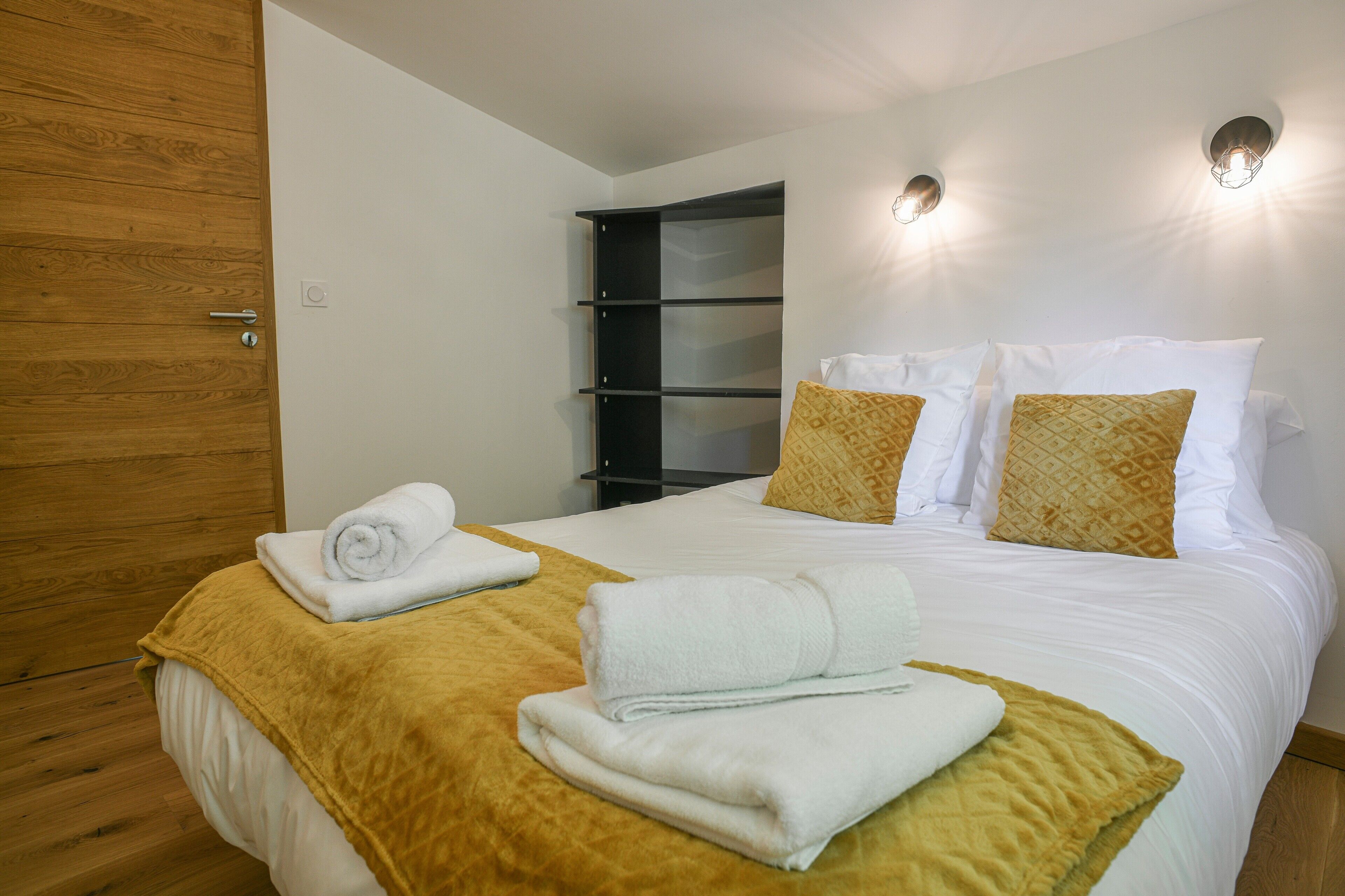 A hotel room with a bed, a wooden wardrobe, a shelf unit, and two wall-mounted lights.