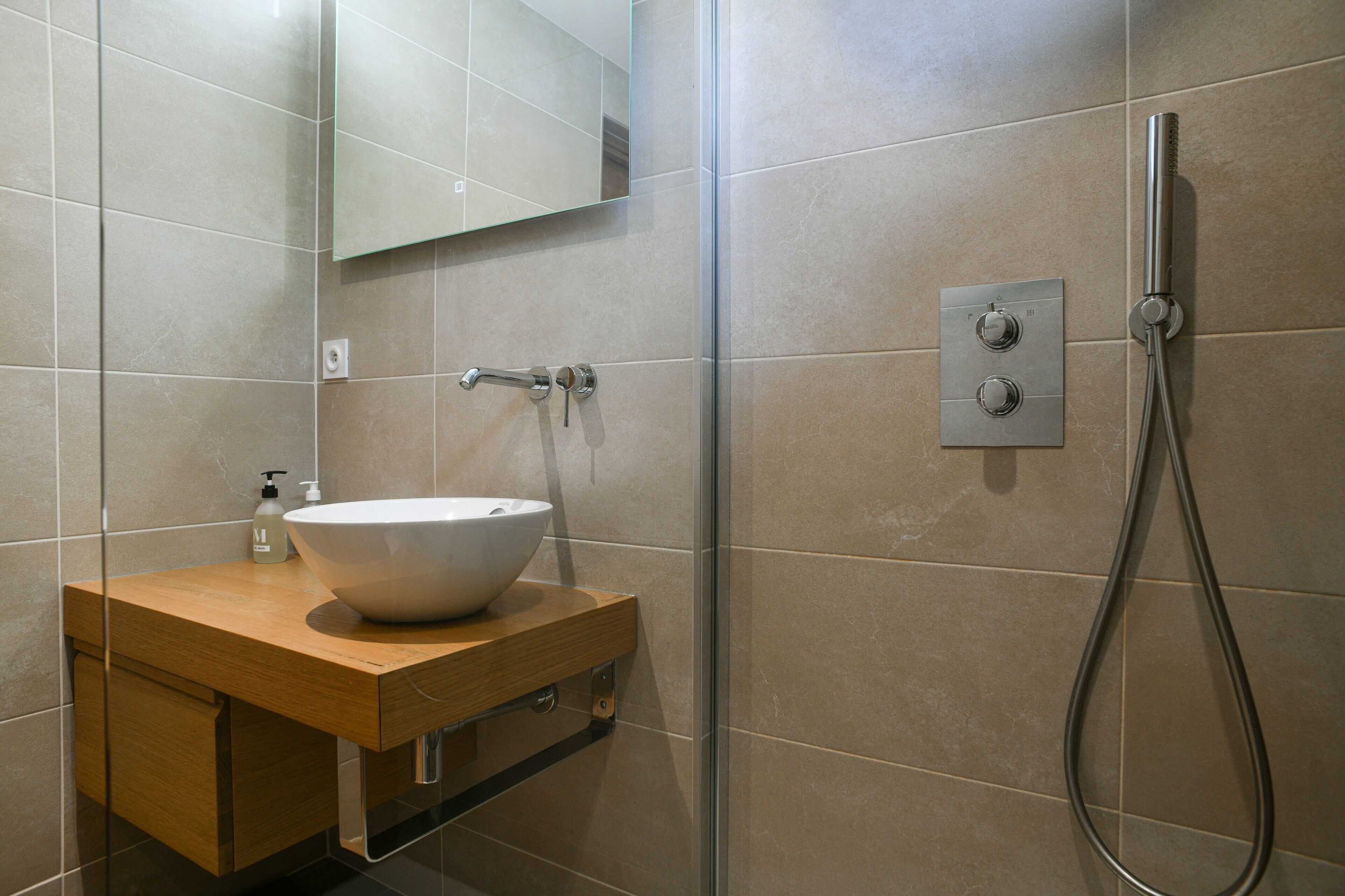 Een badkamer met een houten wastafelmeubel, een witte wasbak, een spiegel en een douche met een handdouche.
