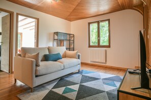 A modern living room with a sofa, a TV, a bookshelf, and a window with a view of greenery.