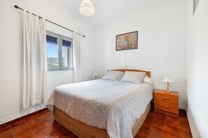 Een slaapkamer met een bed, een nachtkastje, een lamp, een raam met gordijnen en een schilderij aan de muur.