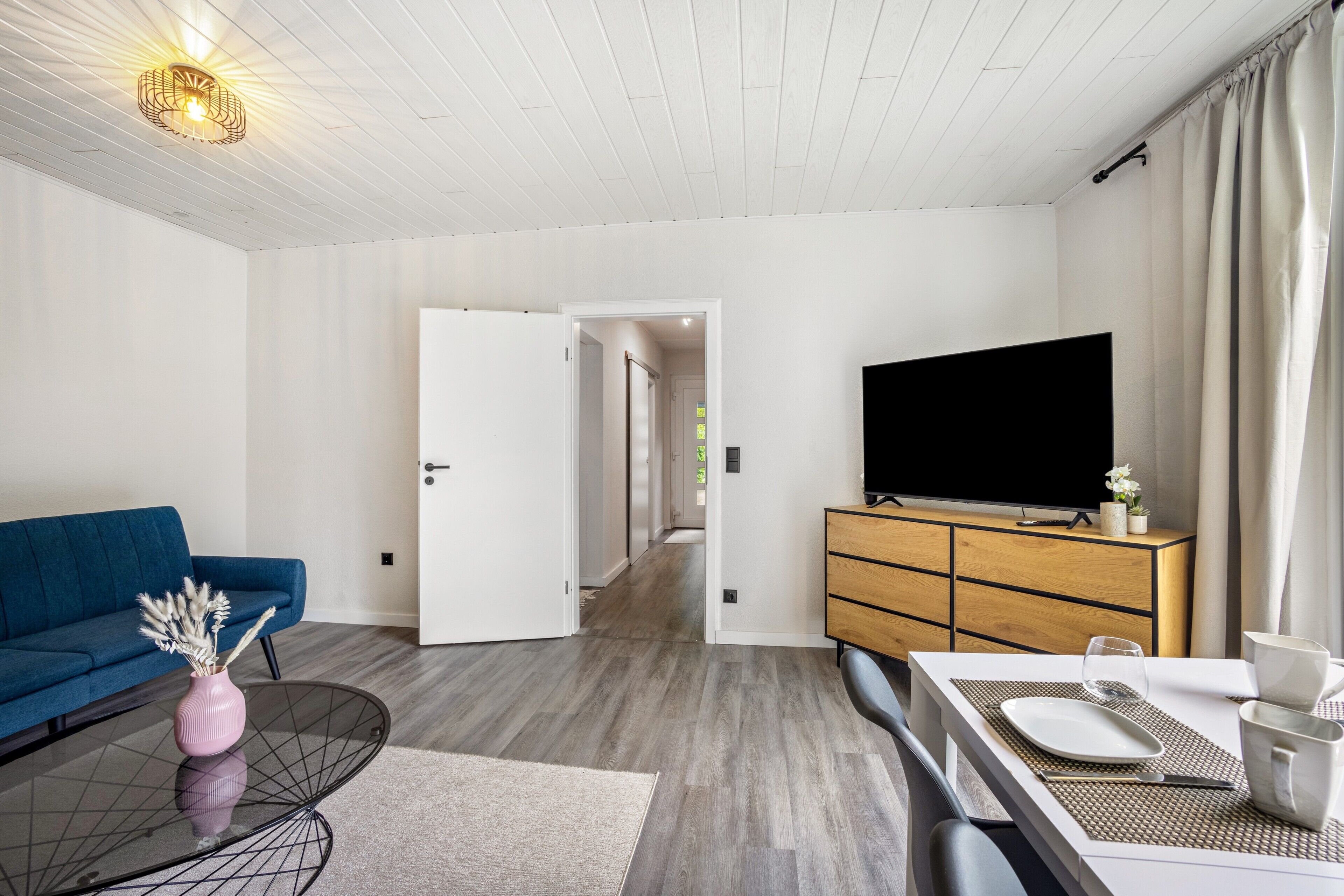 Moderni olohuone, jossa on sohva, kahdelle katettu ruokapöytä, televisio ja puinen kaappi.