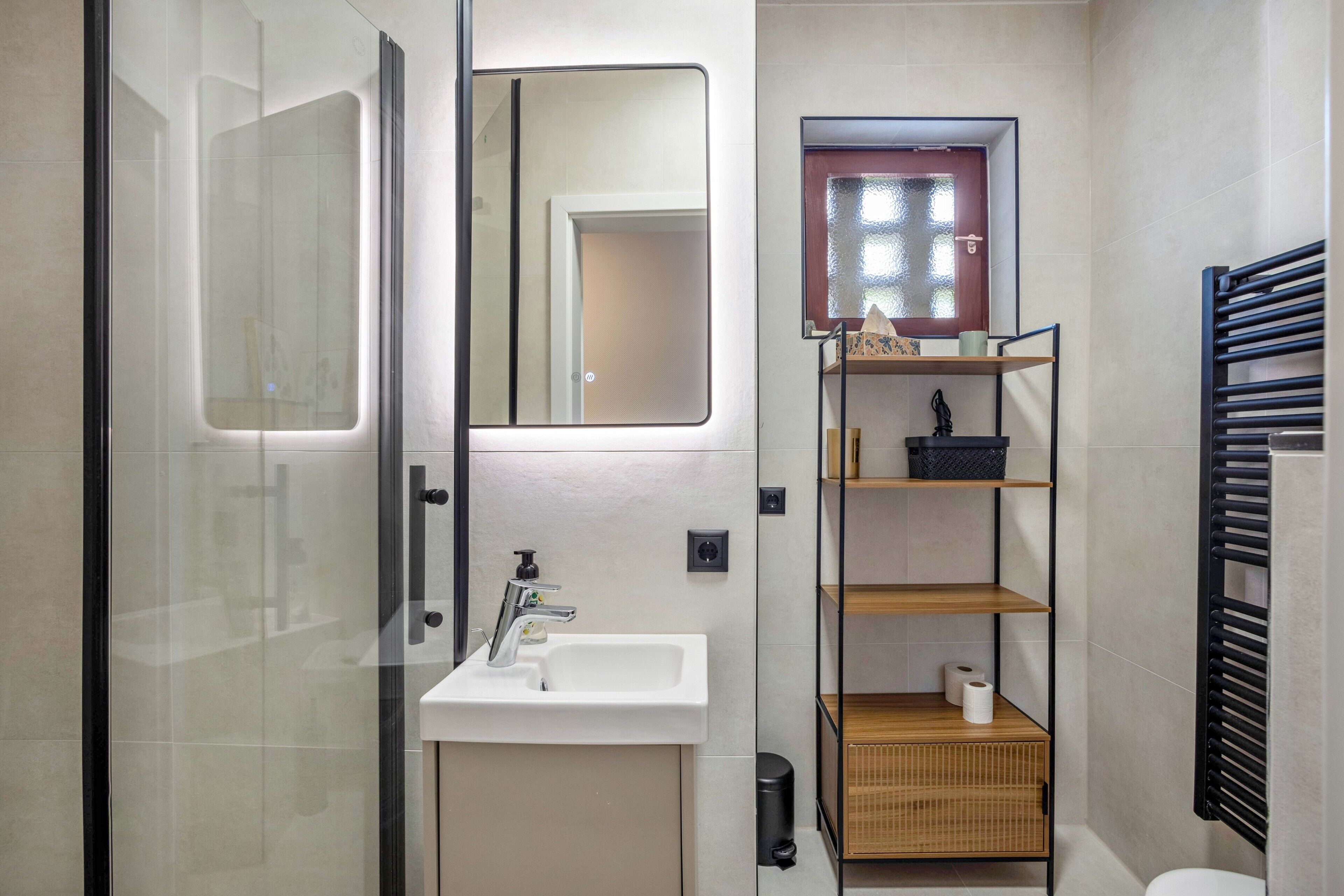 A bathroom with a glass shower enclosure, a white sink, a mirror, a small window, and a black towel rack.