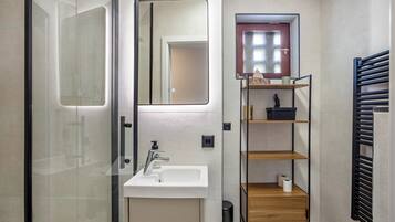 A bathroom with a glass shower enclosure, a white sink, a mirror, a small window, and a black towel rack.