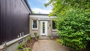 A brick pathway leading to a house with a white door and windows, surrounded by greenery.