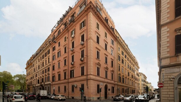 Ein mehrstöckiges Gebäude mit Hauseingang an der Ecke, zahlreichen Fenstern und einer Straße davor.